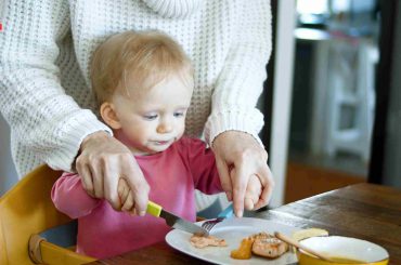 small kid is learning from his mother, the use of knife and fork to eat the food